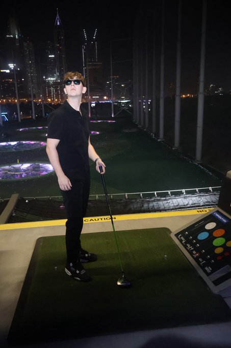 A young man in a black shirt and sunglasses stands on a Topgolf driving range at night, holding a club over a green hitting mat with a "CAUTION" sign visible behind him and the city skyline glowing in the background. The post's initials "GN" likely reference the featured brand **New Era** (NE), seen on the side of his cap, tying the casual streetwear look to the branded headwear in the image.