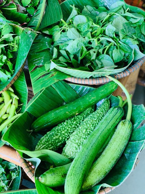 A close-up photograph of fresh green vegetables, including cucumbers and leafy greens, displayed on banana leaves at what appears to be a market.