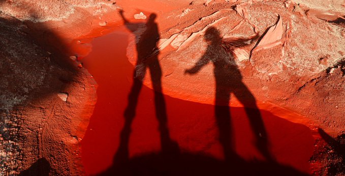 Silhouettes of two people casting long shadows on a reddish rock surface next to a pool of red water, with a third shadow in the foreground.