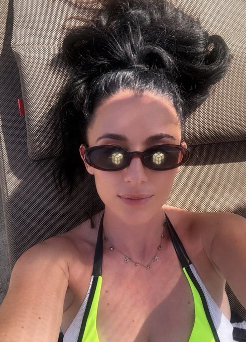 A woman with dark hair styled up lies on a tan lounger wearing slim oval sunglasses and a neon yellow and white bikini, accessorized with a delicate beaded necklace. The image complements the post's vibe of a sun-soaked "GM" (good morning) aesthetic, though no visible text, numbers, or tickers appear in the photo itself to relate to the tokenized private equity theme.