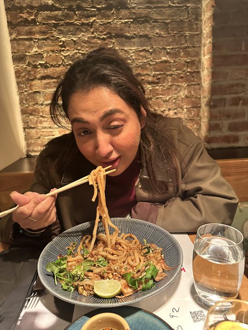 A woman playfully winks at the camera while lifting a tangle of noodles from a bowl of stir-fried udon with cilantro and lime, in front of an exposed brick wall at what appears to be a cozy NYC restaurant. The caption references a throwback dinner in NYC with a friend, matching the intimate dining-out setting of the photo.