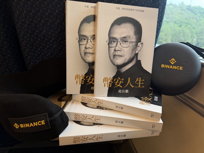 Multiple copies of the Chinese book "币安人生" (Binance Life) by CZ (赵长鹏) are stacked on a tray table next to a Binance-branded neck pillow, with the scenic view visible through the train window during the traveler's journey home.