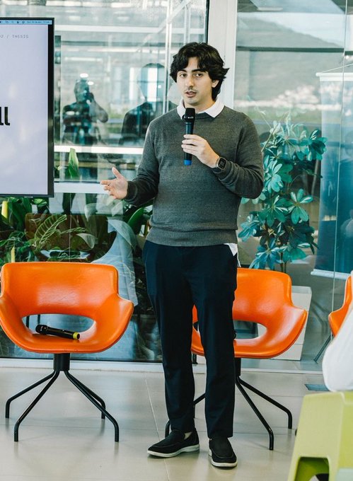 The image features a man standing in front of two orange chairs with his arms outstretched as if giving a speech or presentation. The background includes a large window that allows natural light into the room, creating an inviting atmosphere for the audience. The text on the image reads "Meet our co-founder @luigiremor: A founding engineer at @phase_, Luigi has built core financial systems from zero to production. At Nora, he leads the product and engineering behind $BRS, our Brazilian Real stablecoin and the ecosystem around it."