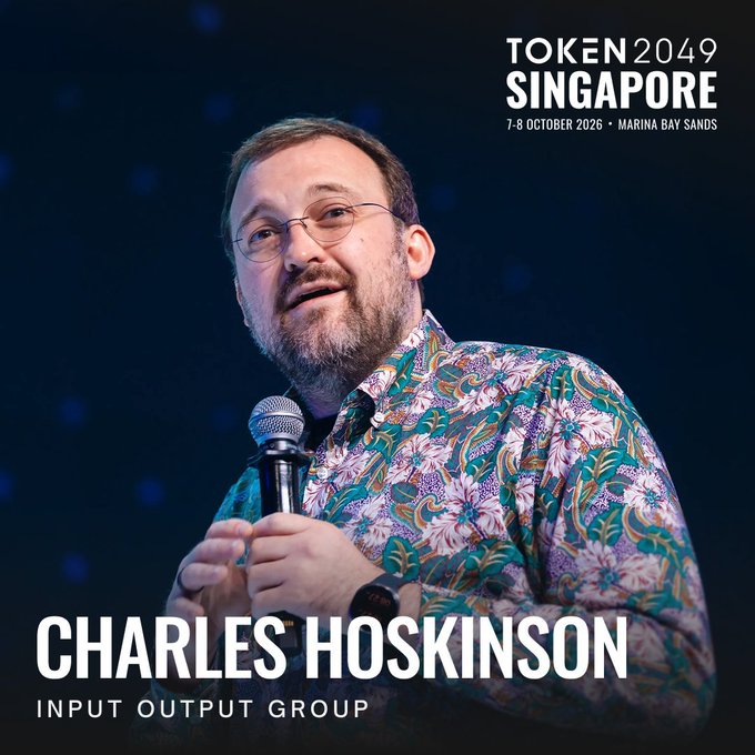 A promotional image for TOKEN 2049 Singapore (7-8 October 2026, Marina Bay Sands) features Charles Hoskinson of Input Output Group speaking into a microphone while wearing a colorful floral shirt.