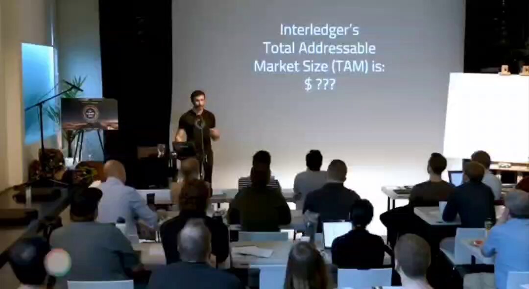 In the image, there is a man giving a presentation to an audience sitting at desks while he speaks into a microphone. The room appears to be quite large with multiple rows of tables, suggesting that this could be a conference or meeting setting. On one wall near the front, there is a projector displaying some information related to Ripple, possibly the current value of XRPLEDGER.