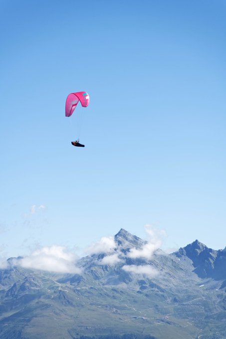 A paraglider with a vibrant pink and blue canopy soars through a clear sky above snow-capped mountain peaks. The distant valley below shows signs of human settlement beneath wispy clouds.