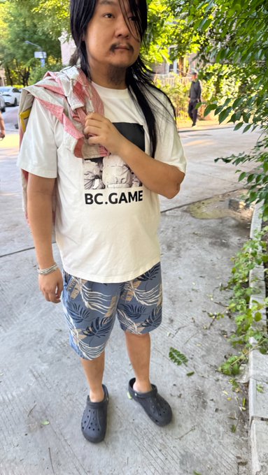 A man stands on a sidewalk wearing a t-shirt featuring "BC.GAME" branding with a towel draped over his shoulder, dressed casually in tropical-print shorts and black Crocs. The image appears unrelated to the post about OKX and OKB crypto sentiment, possibly serving as a personal or lifestyle photo.