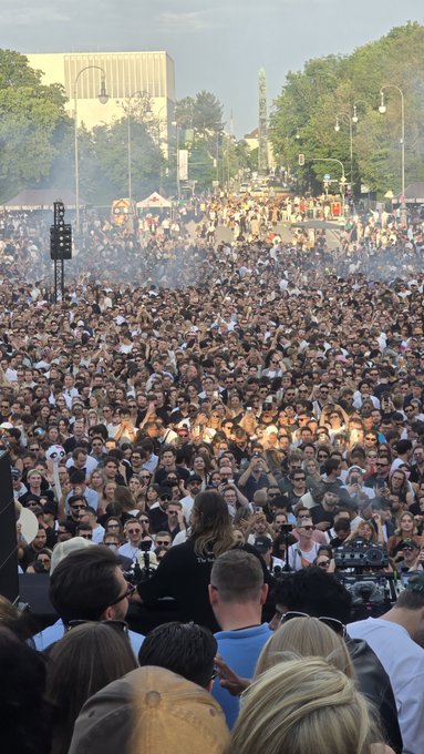 The image shows a large crowd of people gathered together, possibly at an event or concert. There are many individuals standing close to each other, creating a sense of unity among them. The crowd appears to be diverse, with some wearing hats while others do not. In the background, there is a building and trees that add context to the scene. The image also includes a caption that reads "Imagine if they were all SOL maxis." This could possibly be an inside joke or reference to the Solomun concert hall in Paris where the event took place.