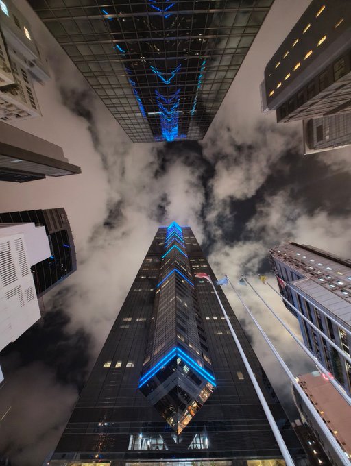 Dramatic low-angle nighttime photograph looking up at skyscrapers, with the central building illuminated by bright blue geometric/chevron light patterns reflecting against the cloudy sky.