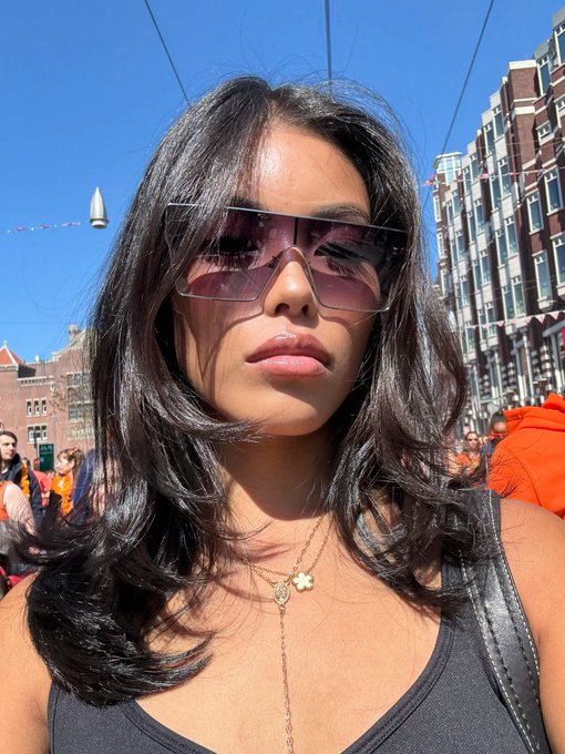 A woman with long dark hair wearing oversized purple-tinted square sunglasses and a black tank top poses for a selfie at what appears to be an outdoor street festival, with a delicate gold necklace and matching lipstick completing her look. The image accompanies a post asking "What's your Monad address?" — likely a crypto/web3 community prompt inviting her to share her wallet address on the Monad blockchain.