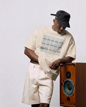 Man wearing a black cap, cream-colored t-shirt with 'SEEN AND YOU WILL FIND' text, and matching shorts, standing next to a wooden speaker cabinet.
