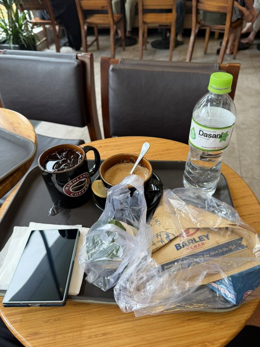 A tray on a table at a cafe containing a mug of iced coffee, a cup of espresso, a bottle of Dasani water, a sandwich in a Bagley Bread plastic bag, and a smartphone.