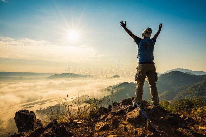 A hiker wearing a backpack and beanie stands triumphantly on a rocky summit with arms raised, overlooking a sea of clouds at sunrise.