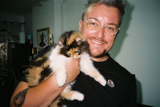 A smiling man with glasses and a beard holds a fluffy calico cat in a warmly lit room with a refrigerator and framed pictures behind him.