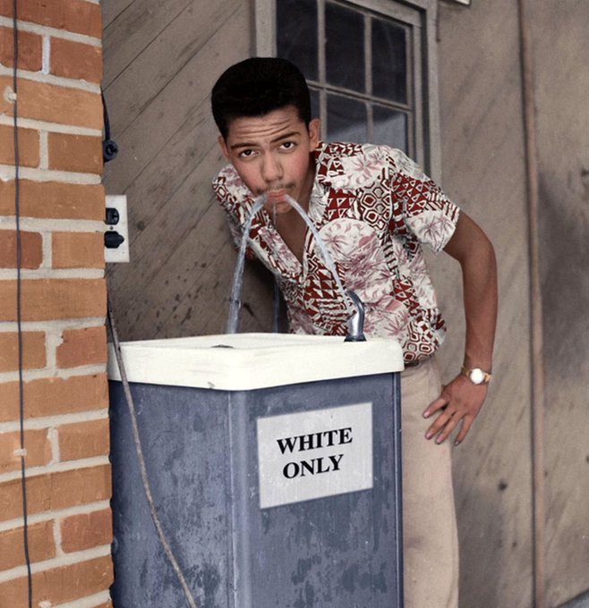 A vintage photograph of a young man in a patterned shirt drinking from a water fountain labeled 'WHITE ONLY'.