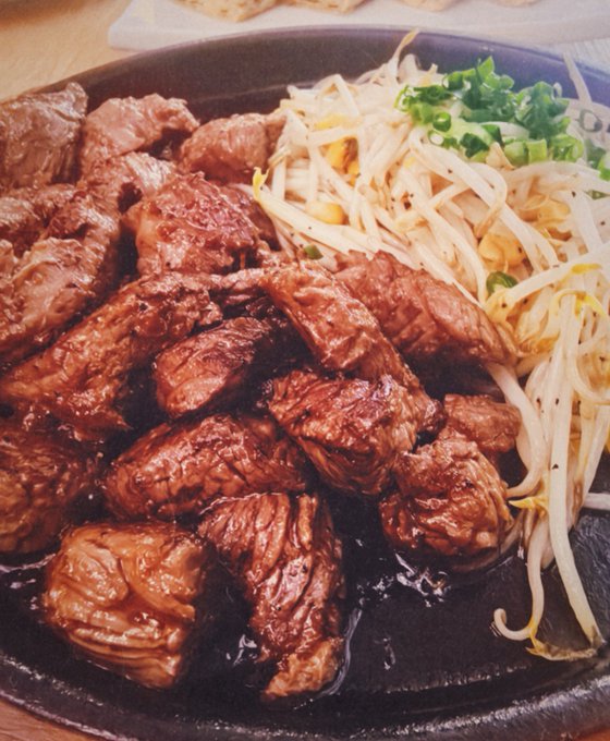 A sizzling cast-iron plate holds cubes of seared steak alongside sautéed bean sprouts topped with chopped scallions, appearing to be a hearty Korean-style beef dish. The caption suggests this is the meal being described in a narrative about a wealthy man's dinner, where he trades his rugged, youthful appearance for a polished luxury brand image while indulging in rich food.