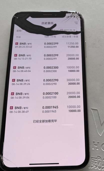 A smartphone with a cracked screen displays a cryptocurrency trading history on a Chinese app, showing multiple BNB/BTC transactions dated August 2017, with text reading '已经全部加载完毕' (All loaded).