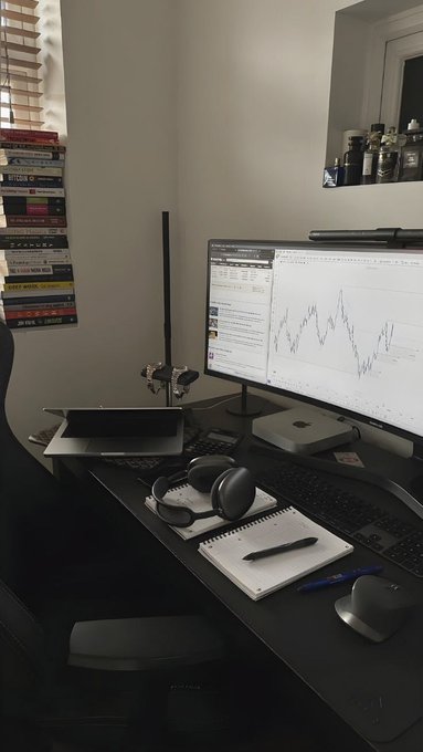 A home office desk setup featuring a curved monitor displaying a stock chart, a laptop, keyboard, mouse, headphones, notebook, and stacked books/DVDs nearby.