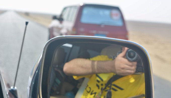 A reflection in a side mirror shows a person wearing a yellow vest holding a camera, with a red vehicle visible ahead on a desert highway.