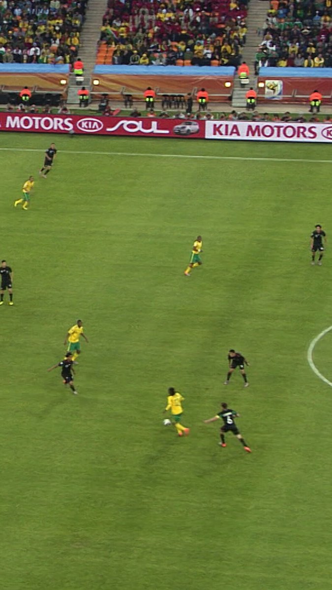 The image shows a group of people playing soccer on a field with a large crowd watching them. The players are spread out across the field, actively engaged in the game. There are at least 13 visible players in various positions, some closer to the foreground while others can be seen further away. A sports ball is located near the center of the image, indicating that it's currently being played with.

The crowd appears to be enjoying the match and cheering for their favorite team or player. The atmosphere seems lively and exciting as everyone is focused on the game.