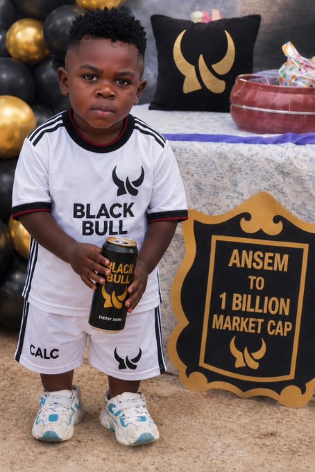 A young child poses at a themed Black Bull celebration, wearing a "CALC" branded shirt and holding a Black Bull "ENERGY DRINK," next to a framed sign reading "ANSEM TO 1 BILLION MARKET CAP"—visualizing the poster's dedication to $ANSEM's journey toward a billion-dollar market cap.