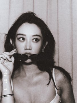 Black-and-white portrait of a young woman with long dark hair lowering her sunglasses to reveal her eyes, with a vertical date stamp on the left.