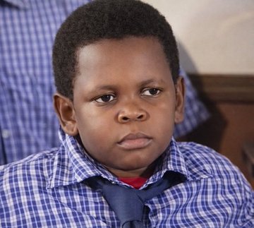 The image features a young boy wearing a blue and white plaid shirt, with his hair styled into a short cut. He is looking off to the side while sitting down. The background of the image shows a person's face, but only their upper body can be seen. There are no texts or other objects in the image.