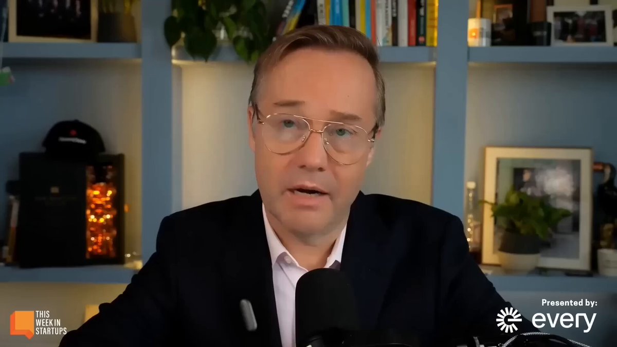 The image shows a man wearing glasses sitting at a desk, talking into a microphone or headset. The background features a bookshelf filled with various items such as books and potted plants. There are also two people visible in the scene, one of whom is holding a camera.