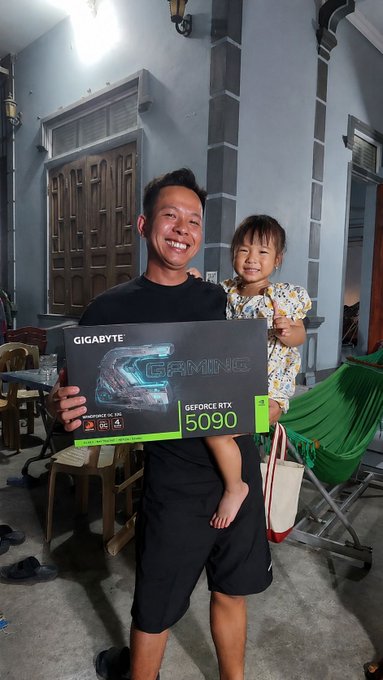 A smiling man holding a child while displaying a Gigabyte GeForce RTX 5090 Gaming OC graphics card box.