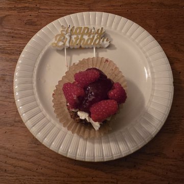 A small dessert topped with fresh raspberries and dark fruit compote, served on a paper liner atop a white plate, with a 'Happy Birthday' decorative pick inserted on top.