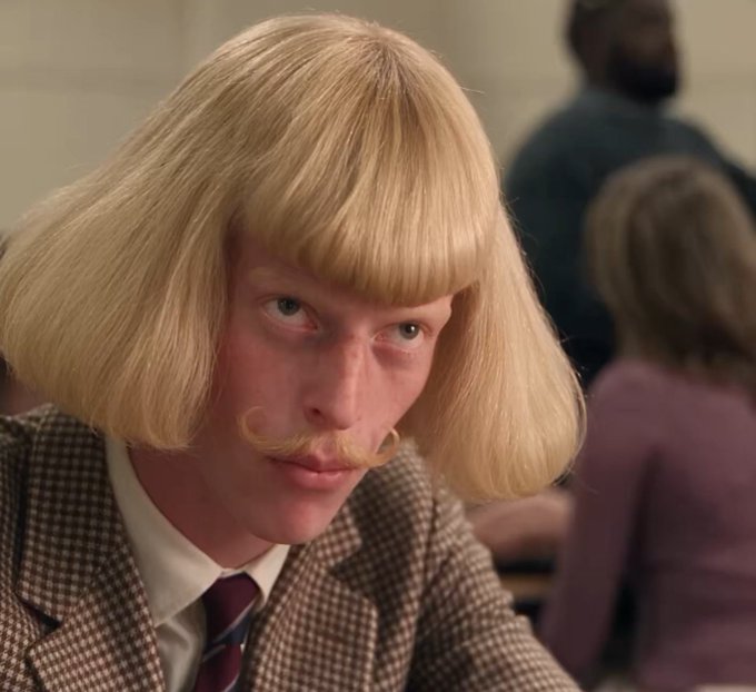 A man with a distinctive blonde bowl-cut wig and curled mustache, wearing a tweed jacket and tie, gazes forward with an intense expression. The image likely references the iconic "Jean-Philippe" character meme, with the post jokingly tying the coin ticker $JEANPHIL to this flamboyant, attention-grabbing persona.
