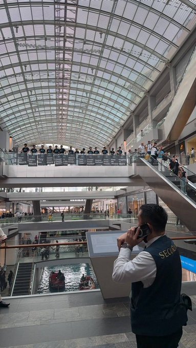 A security guard wearing a vest labeled 'SECUR' stands in a modern shopping mall with a large glass ceiling, talking on his phone while protesters holding banners are visible on the upper level.