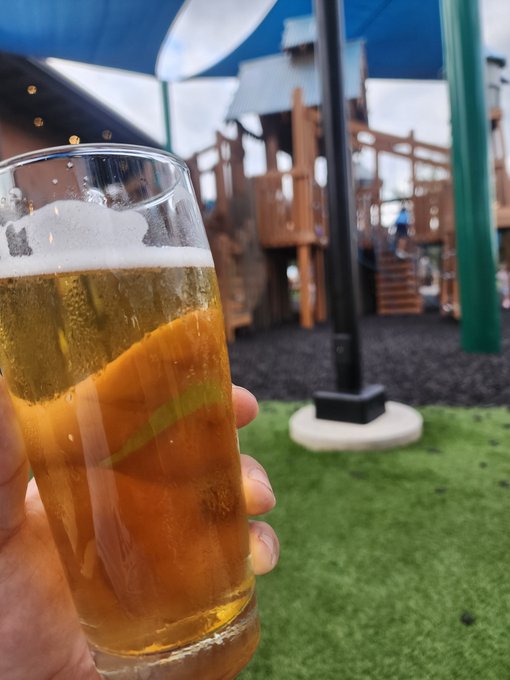 First-person view of a hand holding a glass of beer in front of an outdoor playground area with wooden play structures and green shade sails.