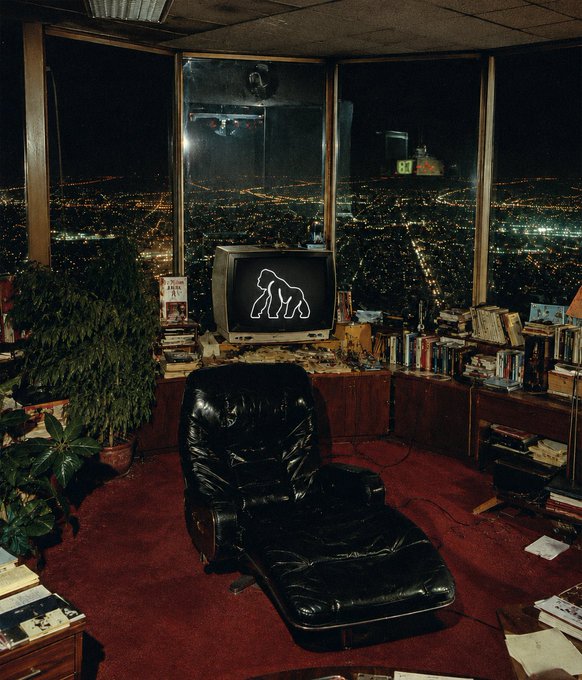 A high-rise penthouse room features a black leather lounge chair facing a vintage TV displaying a glowing gorilla silhouette, with floor-to-ceiling windows overlooking a sprawling nighttime cityscape. The contemplative gorilla imagery and "immaculate vibes" caption tie into the $APE token's primate branding, evoking an air of nocturnal solitude.