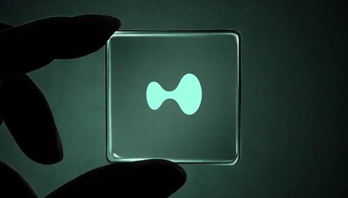 A close-up photograph of a silhouetted hand holding up a small square illuminated panel displaying the glowing teal Hyperliquid logo (a horizontal hourglass shape) against a dark background.