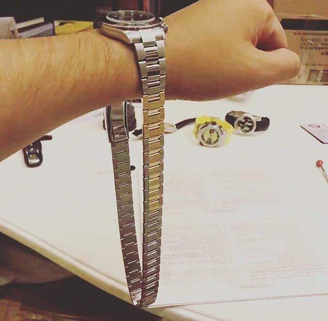 The image shows a person's wrist with a silver watch, which appears to be broken or stopped working. The person is holding the watch up to their face as if trying to fix it. In addition to the watch, there are several other watches on the table and some books scattered around. A red pen can also be seen in the scene.