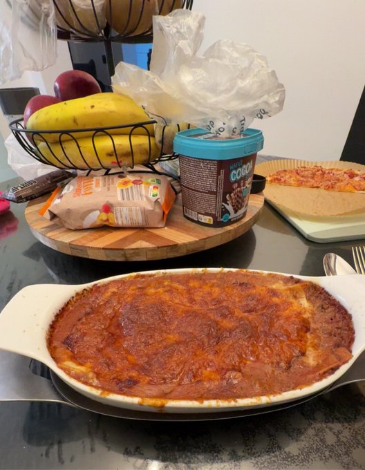 A steaming lasagna dish takes center stage on a dining table, with a wooden cutting board in the background holding a banana, apples, a Cocoa package, and other pantry items. The homemade meal serves as a cozy backdrop to the post's focus on enjoying life while holding $HMM bags and noting good dips on Robinhood.