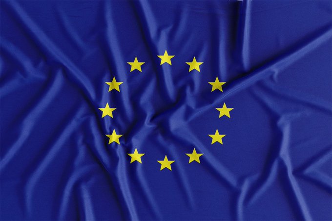 The image features a close-up view of a blue flag with yellow stars, which appears to be waving or fluttering due to its texture. The background is blurred, drawing attention to the flag as the main focus of the scene. In the corner of the image, there are words written in white text that read "European Law Makers Expects Digital €uro To Launch In 2029 And #XRP Is Working With European Central Bank." This information suggests a connection between the flag and the ongoing discussions or developments related to digital currency within Europe.