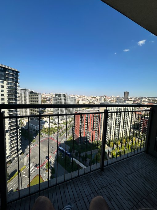 A view from a high-rise balcony looking out over a cityscape with modern apartment buildings and streets under a clear blue sky. A person's legs and feet in striped socks are visible at the bottom of the frame.