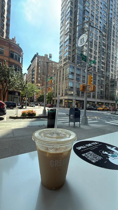 An iced coffee from Gregorys Coffee sits on a table overlooking a sunny Manhattan street corner, perfect for a "gm everyone" morning greeting.