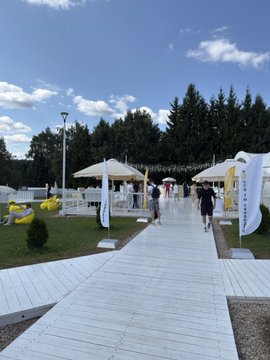 An outdoor event space featuring a paved walkway lined with white canopy tents and decorative yellow flower-shaped installations, set against a backdrop of tall pine trees under a partly cloudy sky.