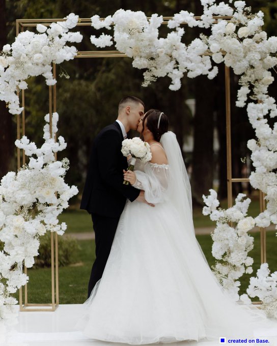 A bride and groom share a kiss under a large, square gold arch decorated with abundant white flowers; a small 'created on Base' logo is in the corner.