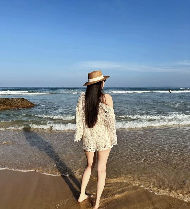 A woman with long dark hair wearing a sun hat and white lace coverup standing barefoot at the edge of the ocean, looking out at the waves.
