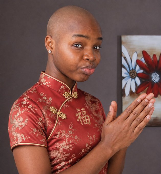 A person with a shaved head wears a traditional red Chinese garment with gold dragon embroidery and a "福" character, posing with hands pressed together in greeting.