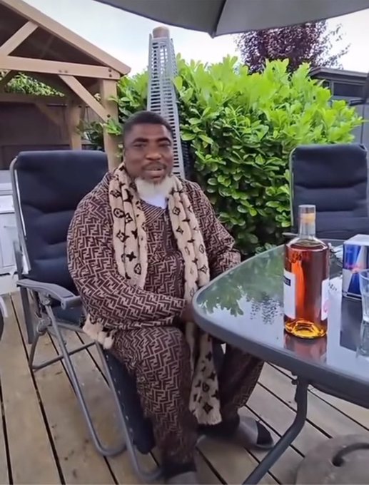 A man wearing a brown patterned robe sits at a patio table with a glass of amber liquor and a Red Bull can, delivering a casual outdoor testimonial about the $TMX booster program on TermMaxFi. The relaxed garden setting reinforces the laid-back, conversational tone of his promotional pitch.