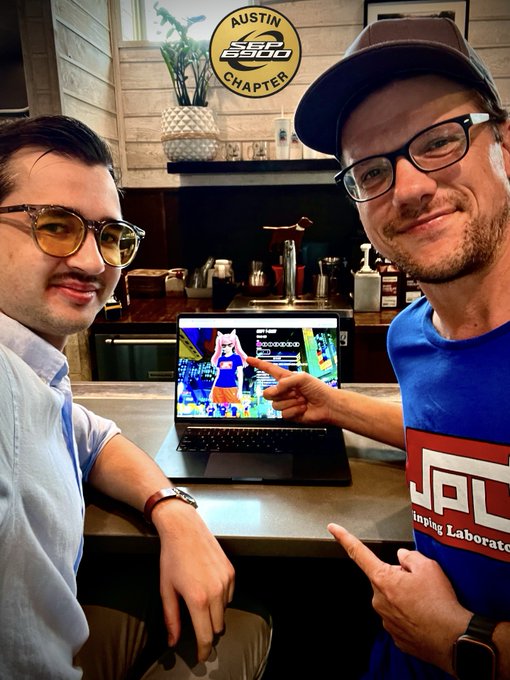 Two men wearing glasses smile for a photo while pointing at a laptop screen displaying an anime character, with an 'AUSTIN SSC CHAPTER' logo visible on the wall behind them.