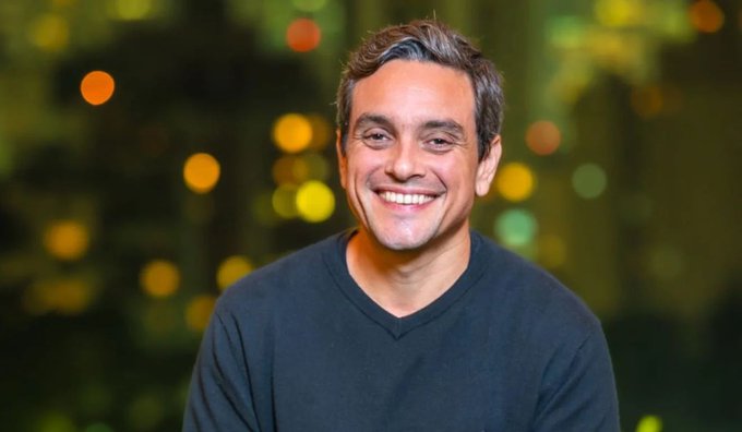 A smiling man with graying hair wearing a black V-neck shirt, photographed against a bokeh background of warm lights.
