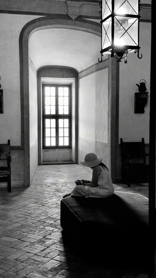 A young girl in a sun hat sits peacefully on a bench in a softly lit, arched corridor, embodying the serene stillness referenced in the fine art piece "Where Peace Rests." An ornate hanging lantern glows above her, complementing the quiet atmosphere of this limited 25-edition Tezos ($XTZ) photograph from the Eirenia collection.