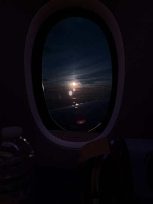 A view through an airplane window at night reveals a dark sky with distant lights and what appears to be the sun or moon glowing on the horizon, suggesting the traveler is en route to their destination. This fits with the post about heading to Japan for Blockchain Week, capturing the journey before arriving to speak at the event.