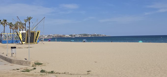 A sunny beach scene with sand, palm trees, a yellow beach structure, and a view of the ocean and distant coastline.